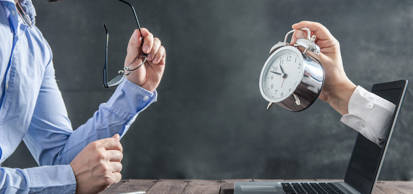 Businessman is sitting at the desk and is looking at the hand with clock coming out of the laptop. Metaphor of spending time at work